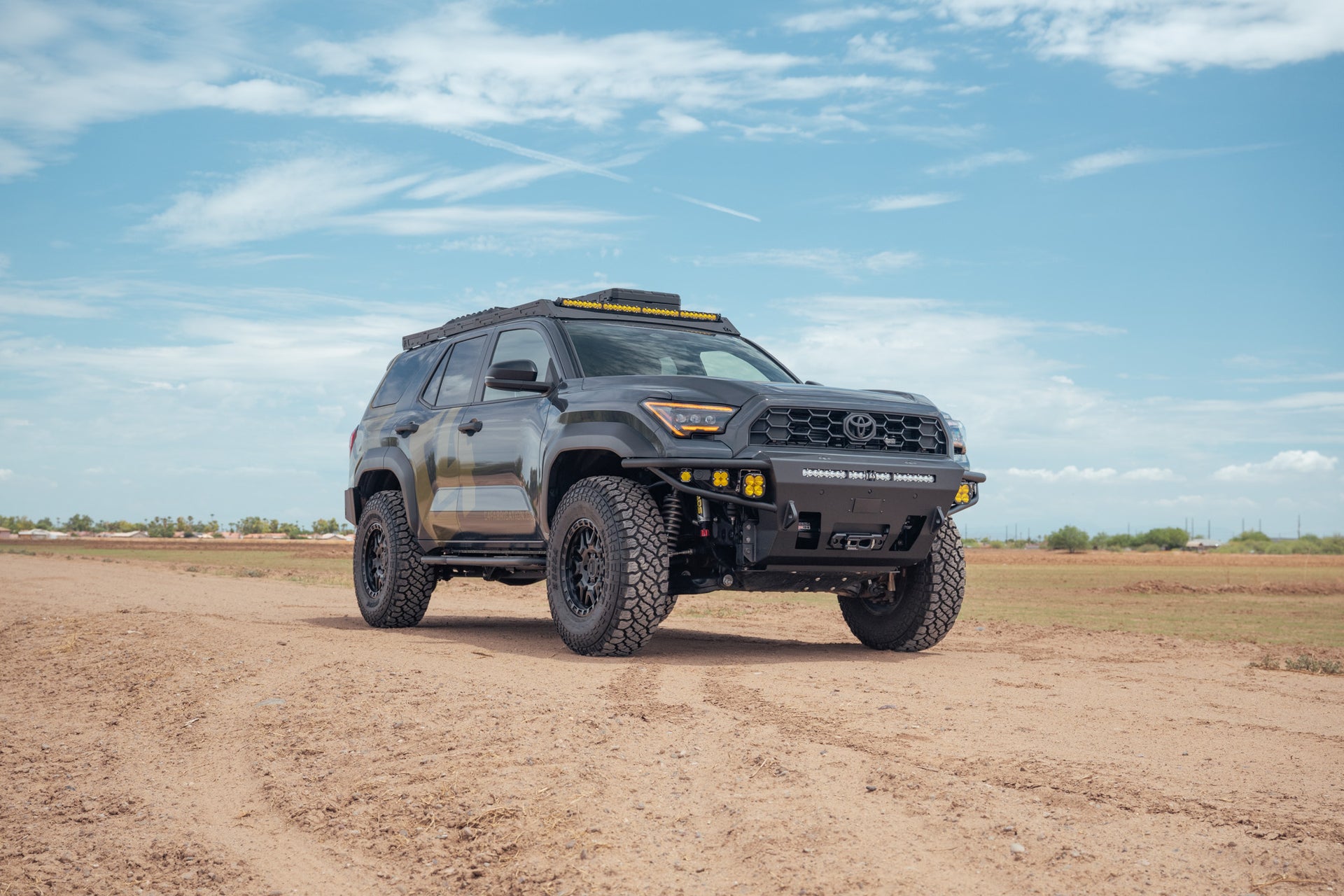 A black Toyota 4Runner SUV with off-road modifications, including a C4 roof rack with light bar and C4 Hybrid front bumper, driving on a dirt road under a blue sky.
