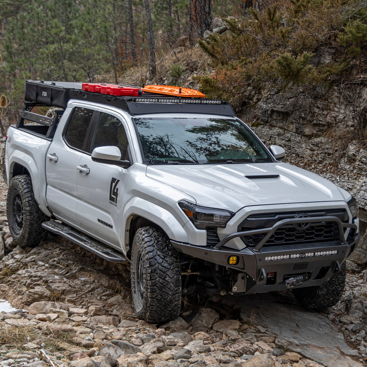 Modified 2024 Toyota Tacoma navigating a rocky stream bed and equipped with a C4 Overland series winch bumper.