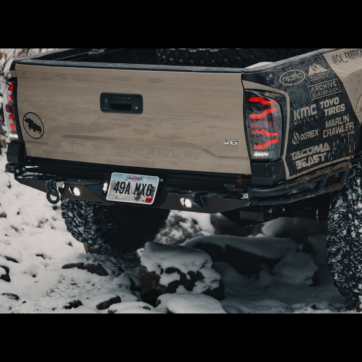 Rear quartering view showing an installed C4 Rock Runner bumper on a tan Tacoma wheeling in the snow.