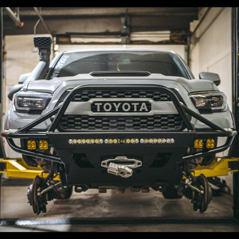 White Tacoma on a lift with the wheels off and zoomed in on the C4 Hybrid winch bumper with full compliment of lighting.